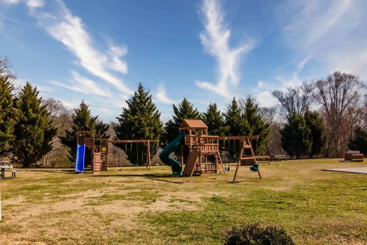A cheerful playground scene with swings, a slide, green grass, and trees, showing children’s play areas and space for families to relax and enjoy the outdoors.