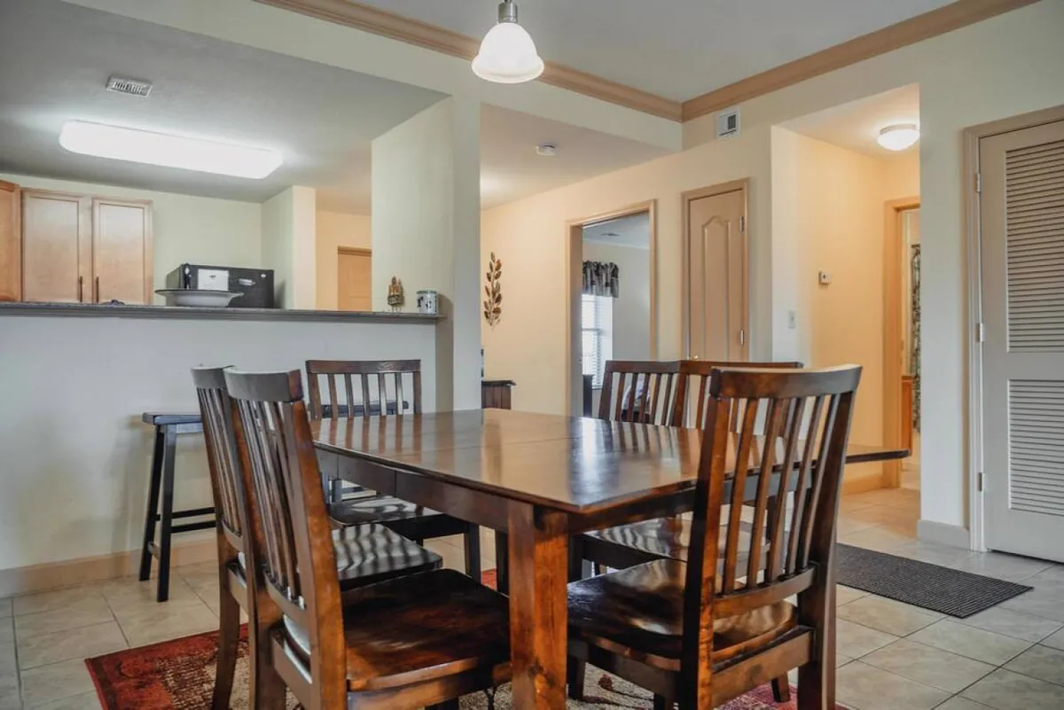 A cozy dining area with a wooden table, matching chairs, and the kitchen counter visible in the background. The scene suggests a space designed for shared meals, celebrations, and casual gatherings with natural light adding to the welcoming feel.