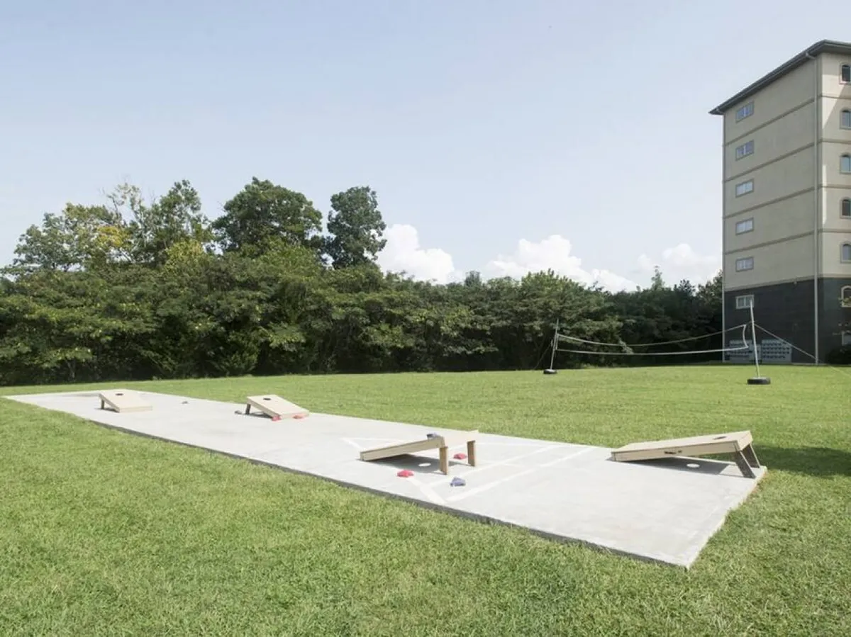 A playful outdoor scene featuring cornhole boards on a grassy area, surrounded by trees and a glimpse of the building in the background. The area invites outdoor games, gatherings, and family fun in a natural setting.