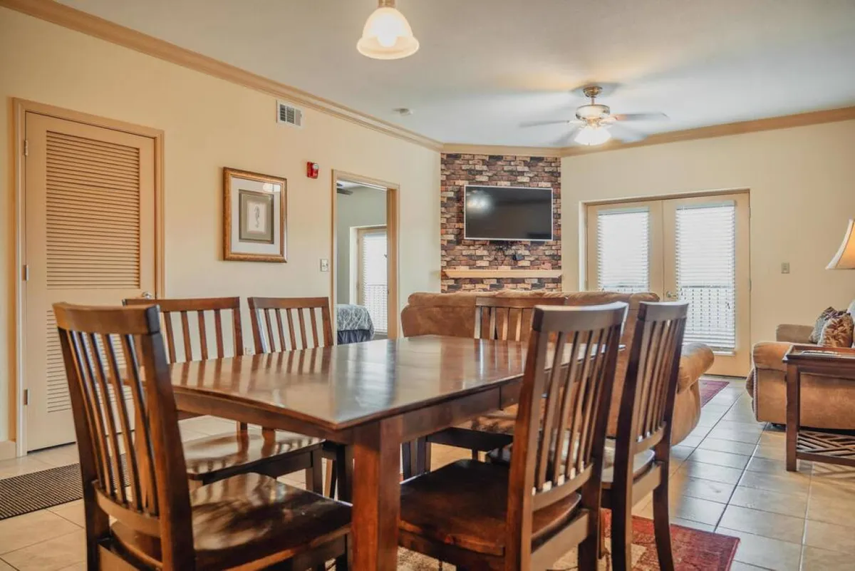 A welcoming, open-concept living area featuring a stone accent wall, a comfortable couch, a large flat screen TV, and a dining table set for group gatherings. The space is accented by warm lighting and decorative touches, making it ideal for family dinners, movie nights, and relaxed conversations.