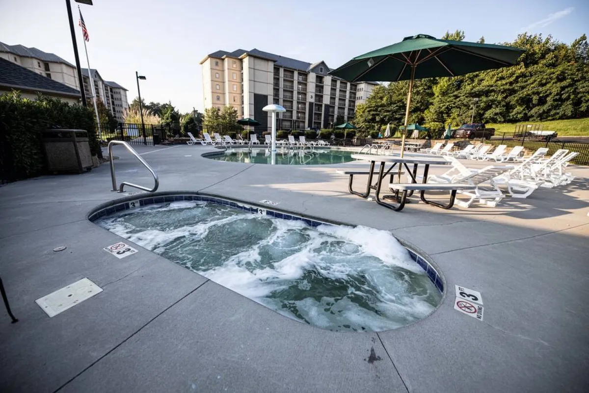 The image showcases the resort's inviting outdoor amenities featuring a sparkling swimming pool, hot tub, lounge chairs, picnic table, and sun umbrella. The area is surrounded by greenery, highlighting space for leisure and socializing.