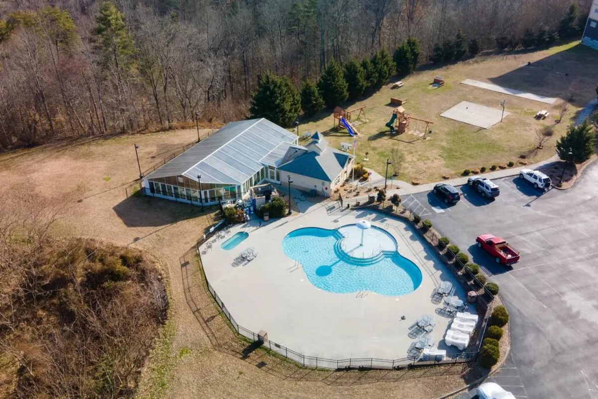 An exterior shot showing the resort's outdoor pool, a clubhouse, a playground area, and a parking lot. The scene is lively, with open spaces perfect for group activities and family gatherings.