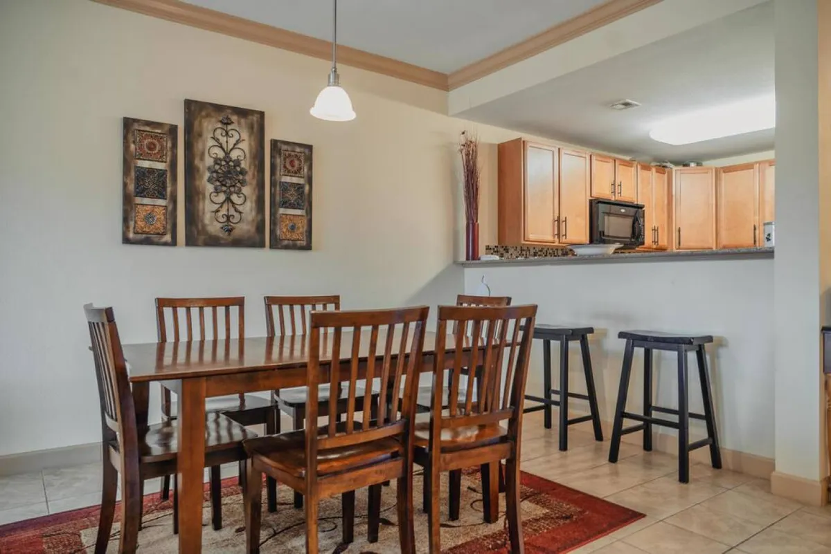 A bright dining area featuring a wooden table with matching chairs, decorative wall art, and a kitchen bar area in the background. The setting is inviting and encourages conversation over meals or games, seamlessly connecting to the kitchen for easy interaction.