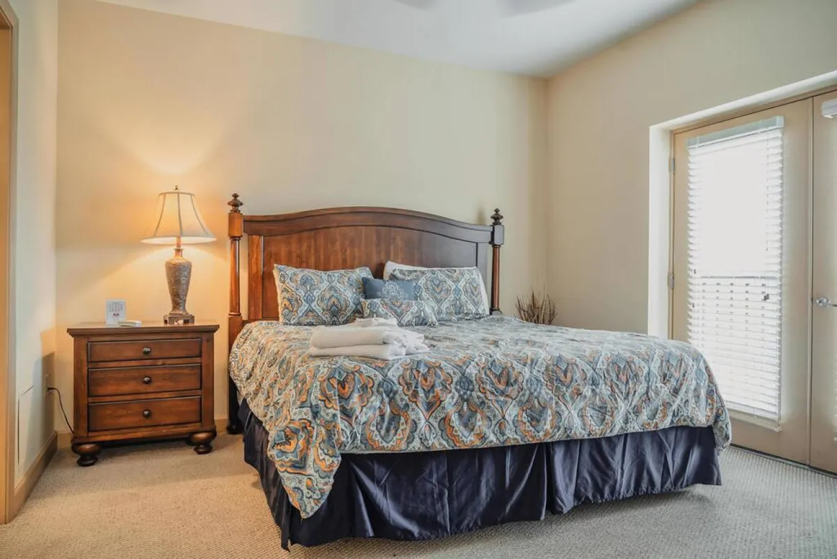 A beautifully appointed bedroom with a plush bed dressed in crisp linens, a warm bedside lamp casting a gentle glow, and a large window letting in soft morning sunlight—creating an inviting and tranquil retreat.